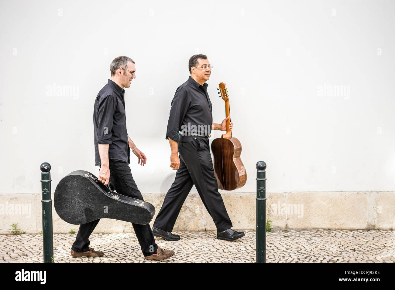 Two guitarists walking with their instruments on the pavement by the white wall Stock Photo