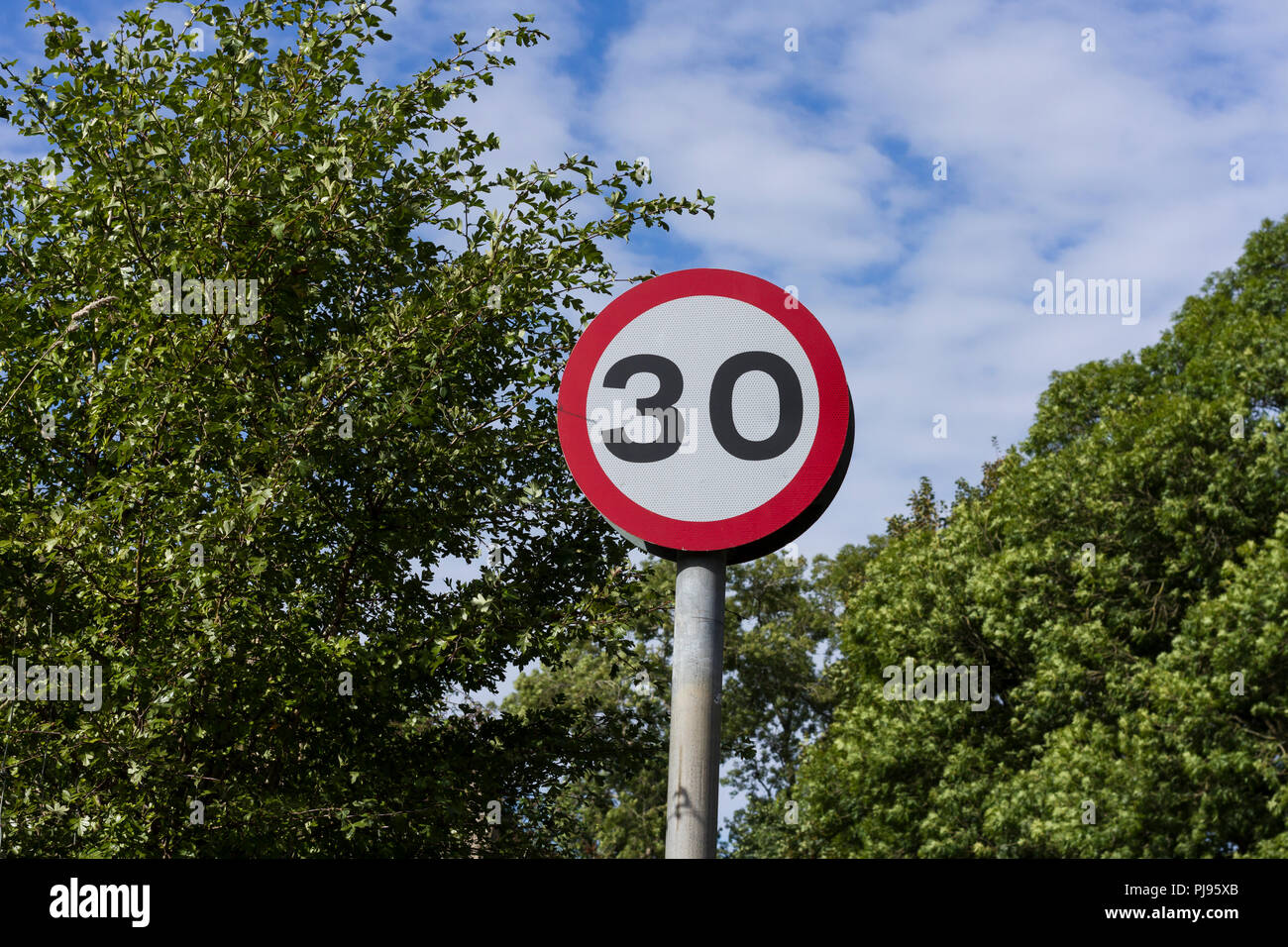 30 mph, round, red & white British road traffic sign against trees & blue sky with clouds background, England, UK Stock Photo