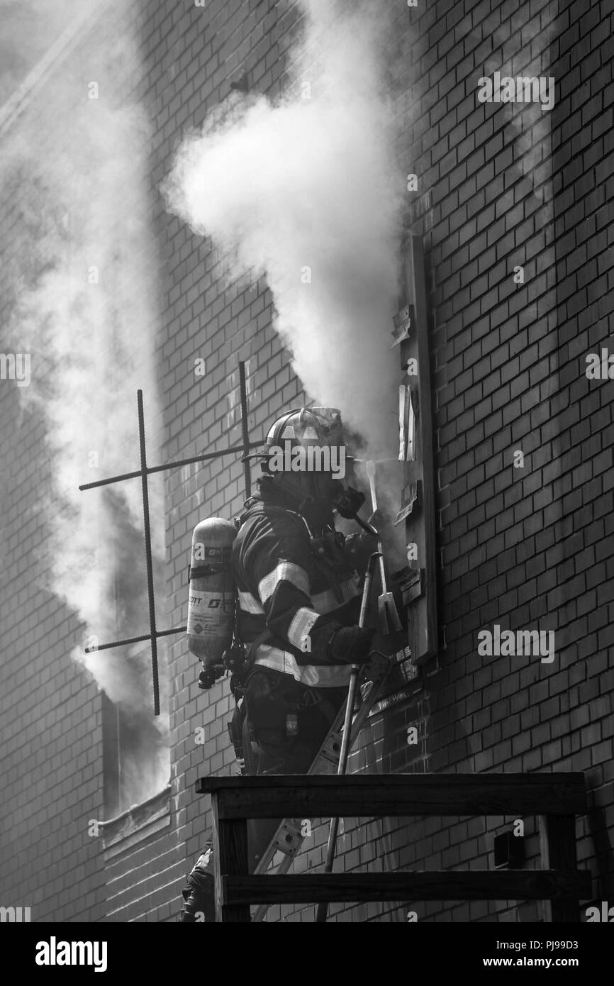 A firefighter prepares to enter a window of a burning training building at The New York City Fire Department Academy on Randall's Island, New York City, New York, on July 9, 2018. New York City Fire Department (FDNY) with U.S. Army North, and U.S. Northern Command (NORTHCOM) conducts training as a joint exercise simulating Chemical, Biological, Radiological, and Nuclear (CBRN) events to maximize the appropriate  response from first responders. Stock Photo