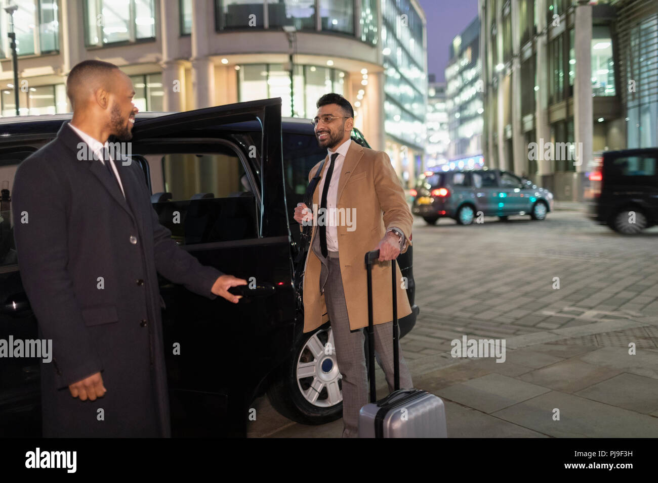 Driver opening car door for businessman with suitcase on urban street at night Stock Photo
