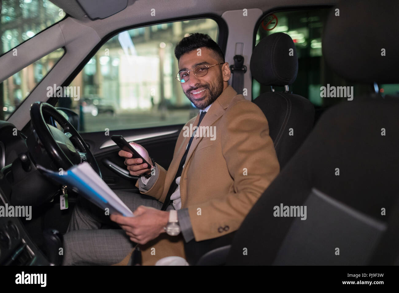 Portrait smiling businessman with smart phone reading paperwork in car at night Stock Photo