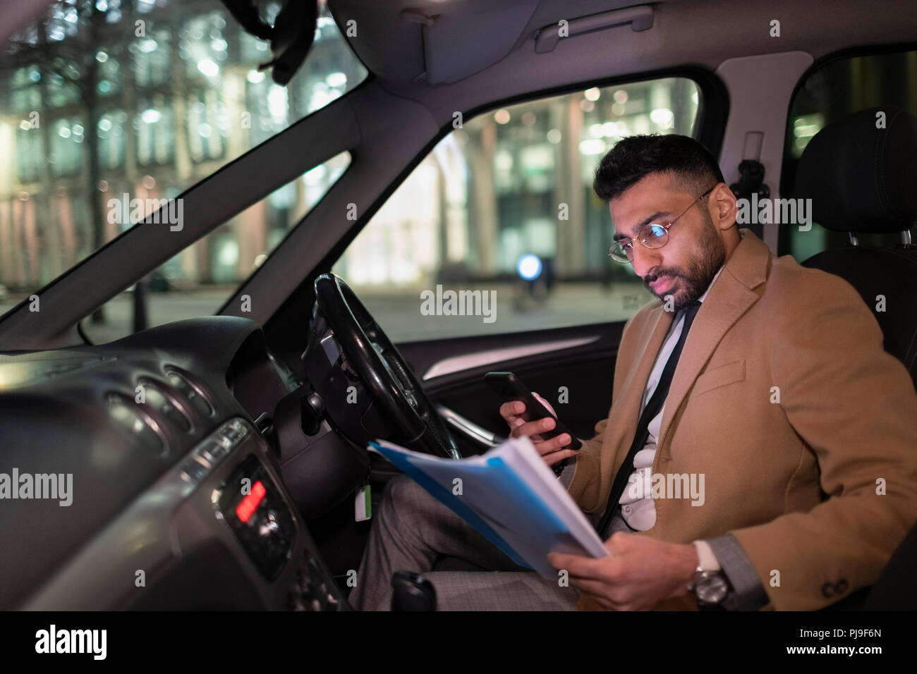 Businessman with smart phone reading paperwork in car at night Stock Photo