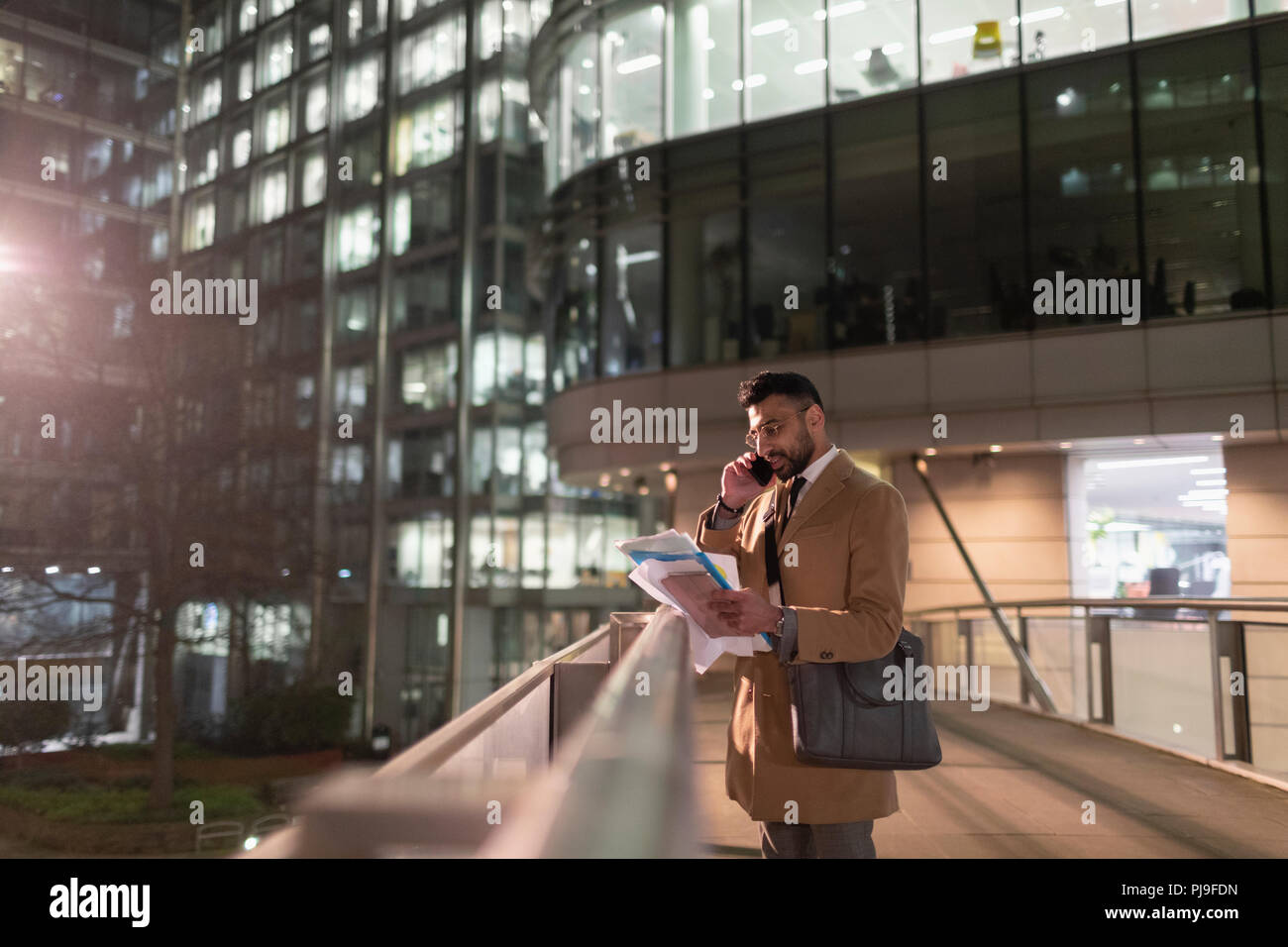 Businessman talking on smart phone and reading paperwork on urban pedestrian bridge at night Stock Photo