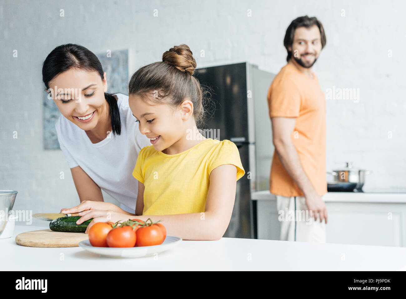mother and daughter preparing salad while father standing blurred on background at kitchen Stock Photo