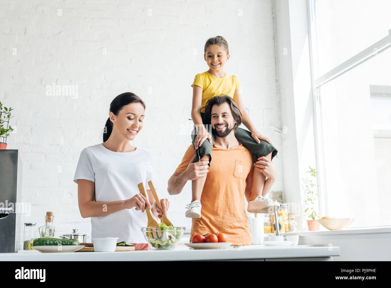 smiling young woman preparing salad while her daughter riding on shoulders of husband at kitchen Stock Photo