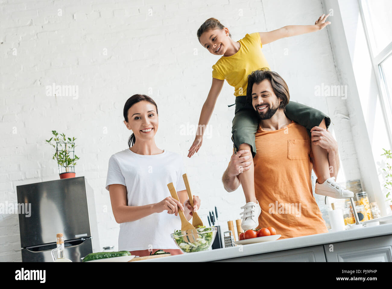 happy young woman preparing salad while her daughter riding on shoulders of husband at kitchen Stock Photo