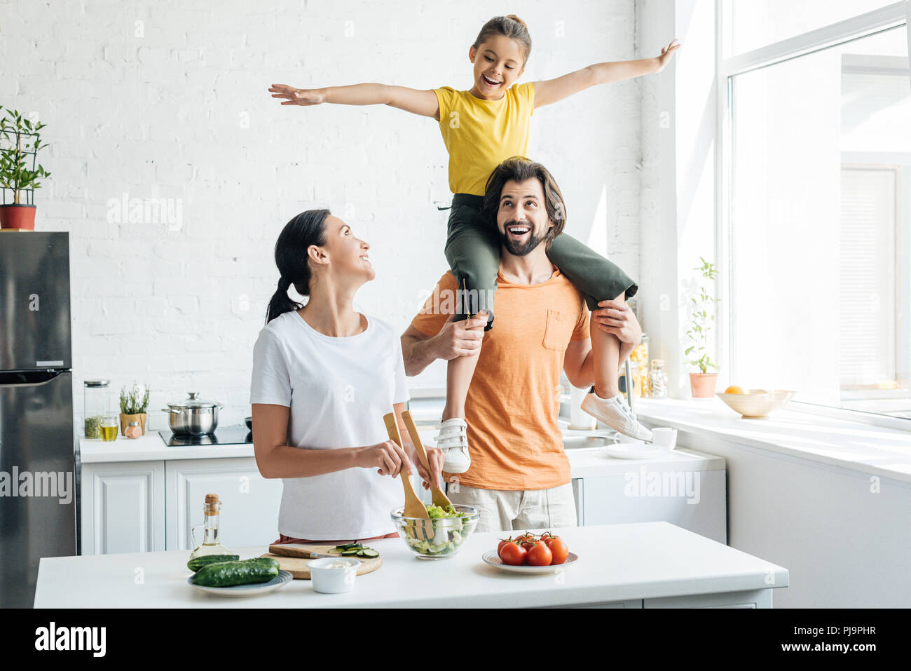 beautiful young woman preparing salad while her daughter riding on shoulders of husband at kitchen Stock Photo
