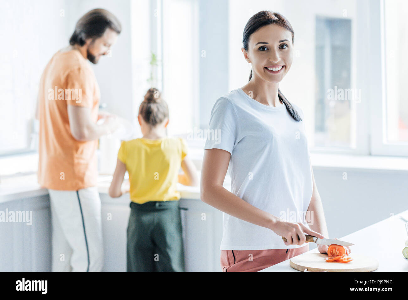 beautiful young woman cutting tomato for salad while her daughter and husband standing blurred on background Stock Photo