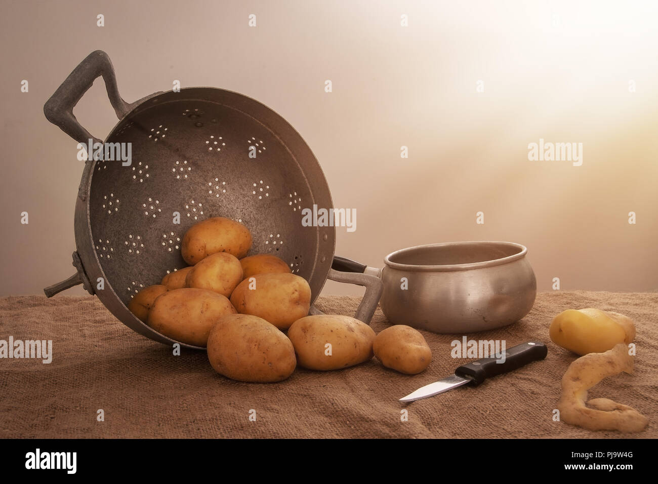 Food preparation, peeled potatoes in farmhouse rustic setting still life with saucepan, collander, knife, hessian aka jute. Stock Photo