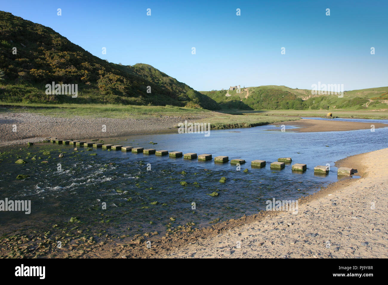 Stepping stones cross the stream at Three Cliffs Bay on the Gower peninsula near Swansea, Wles, UK, with Pennard Castle on the hillside in the backgro Stock Photo