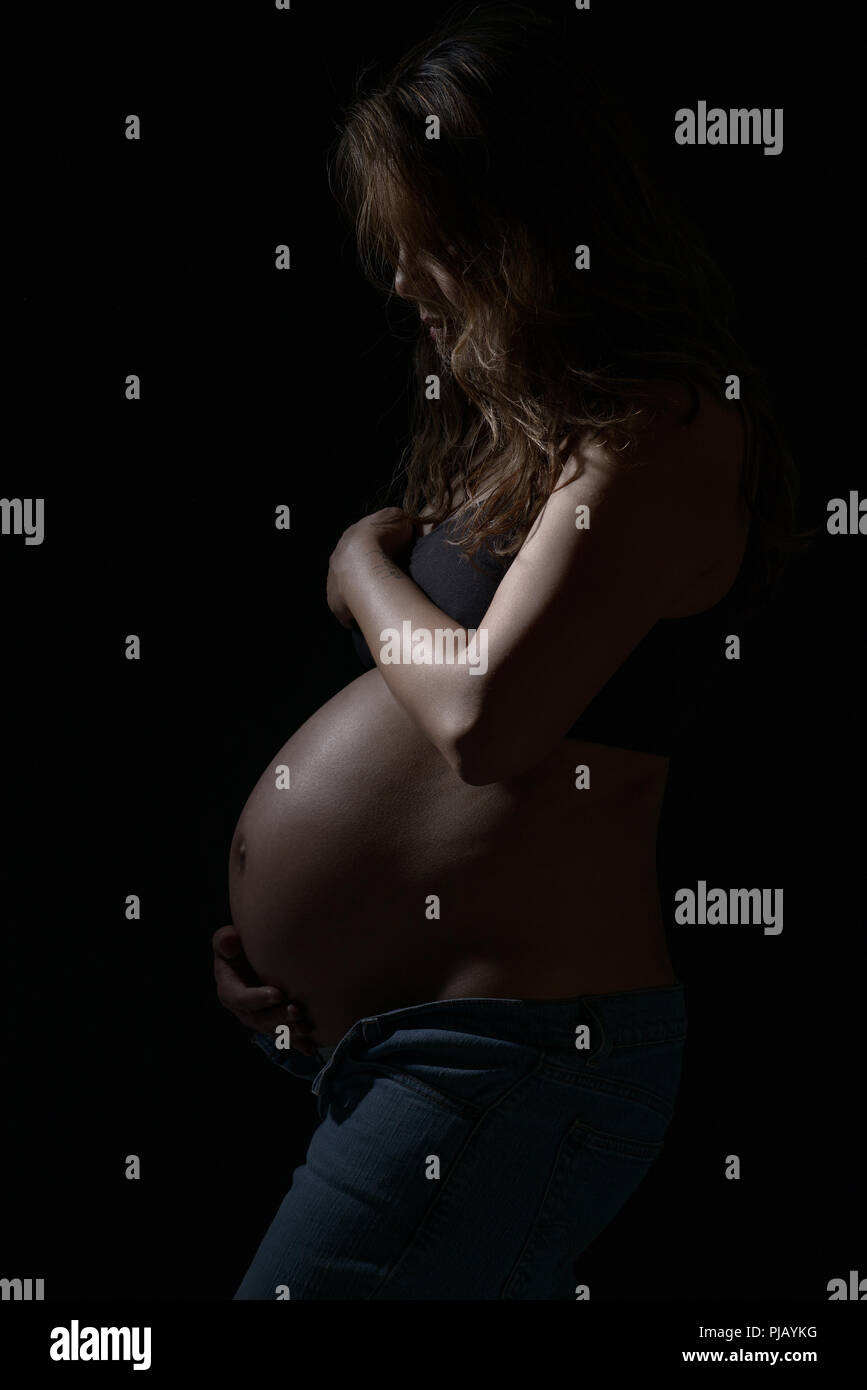 A pregnant Asian woman posing to the side, looking down. A dark artistic portrait. Stock Photo