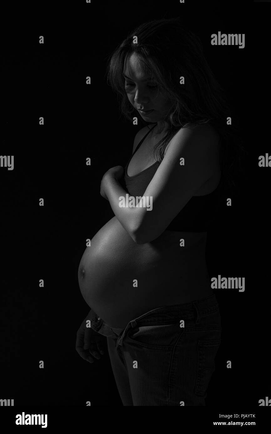 A pregnant Asian woman posing to the side, looking down. A dark artistic black and white portrait. Stock Photo