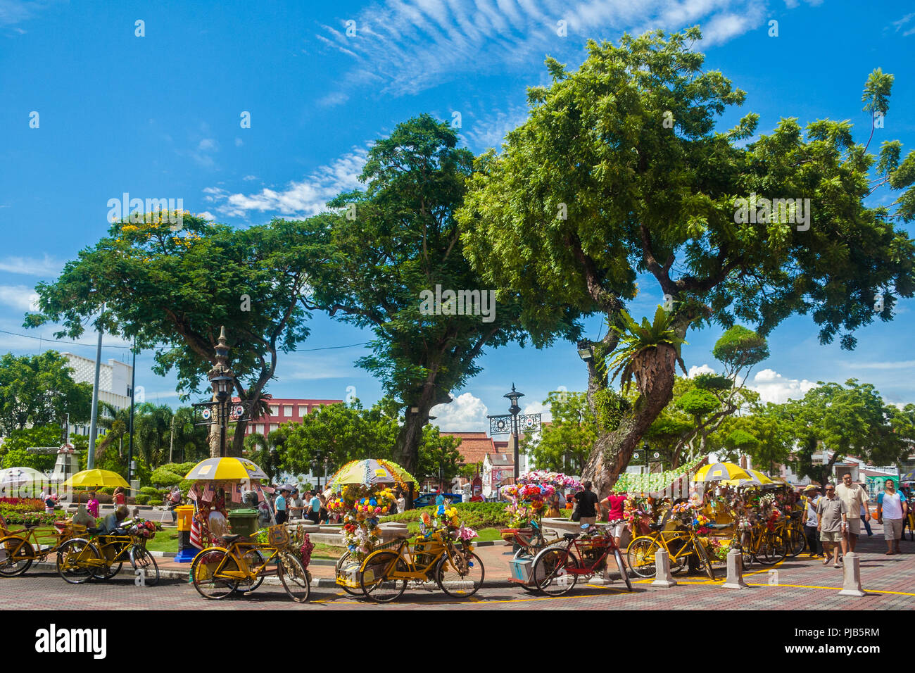 Tourists strolling along nicely decorated yellow trishaws near the fountain at the Dutch Square (Stadthuys area) under trees shade on a beautiful... Stock Photo