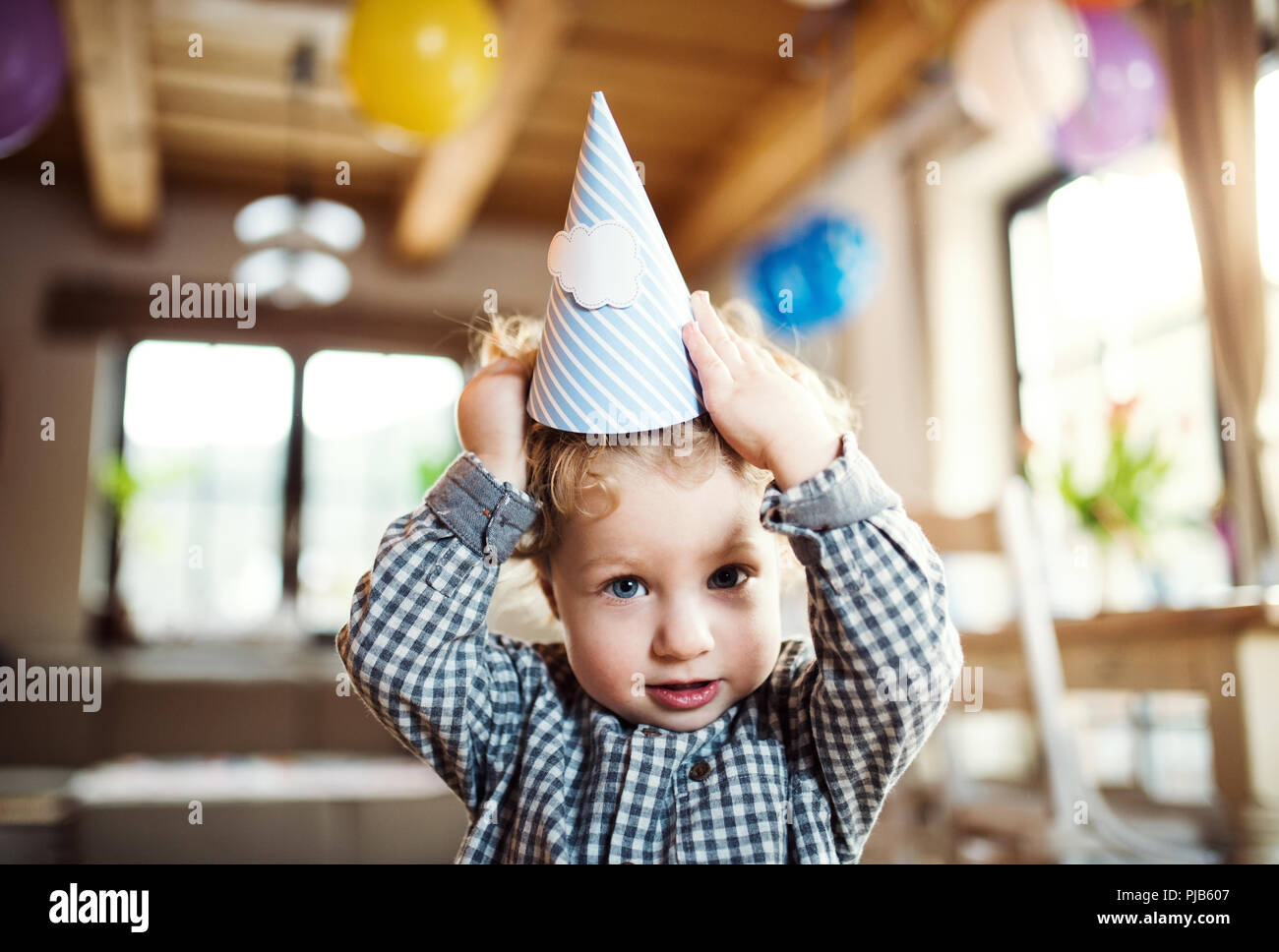 A toddler boy with a party hat standing inside at home. Stock Photo