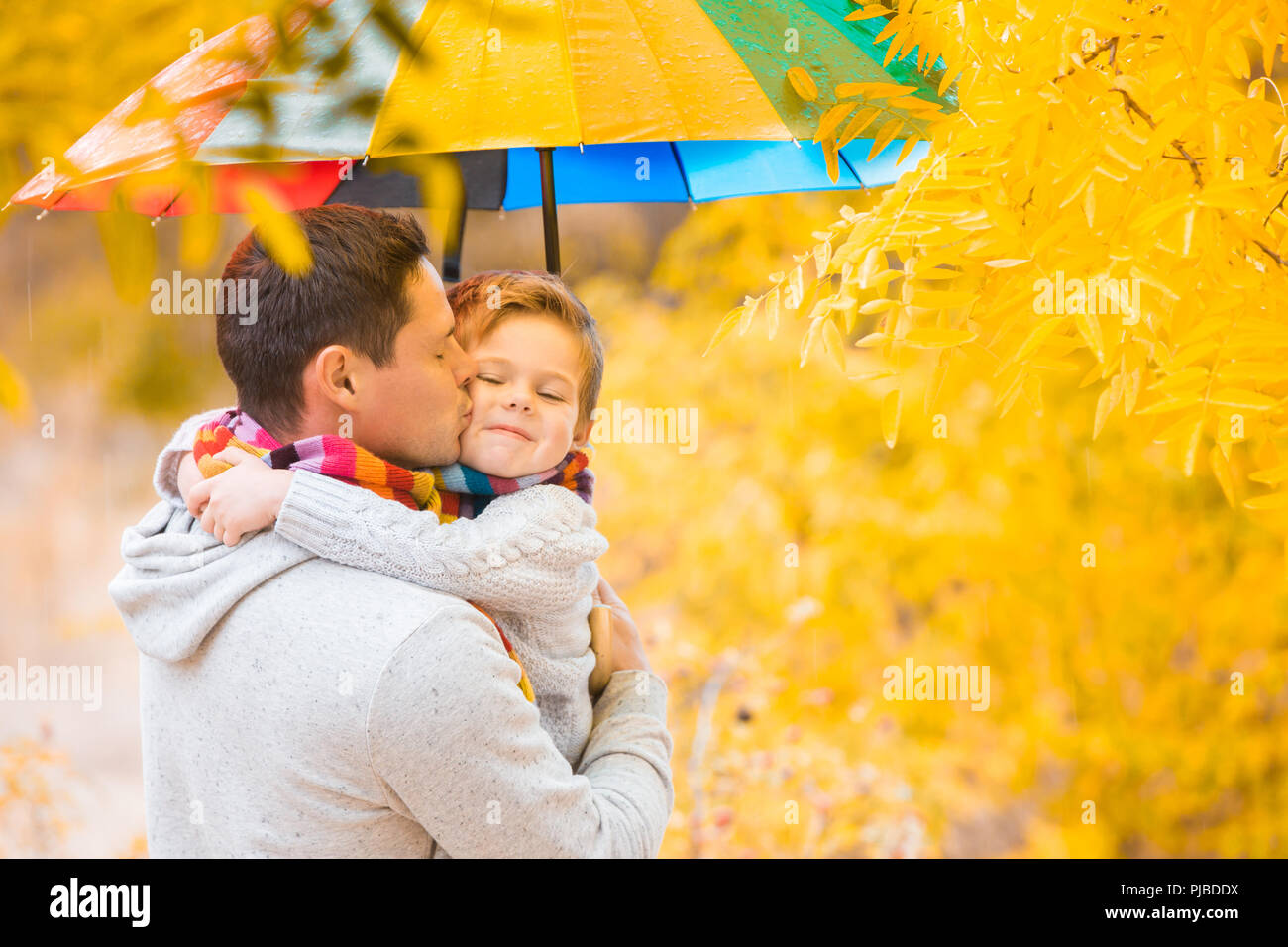A little boy and his father are under a colorful umbrella. Family in warm clothes and colorful scarves. the kid catches the rain drops with his palm Stock Photo