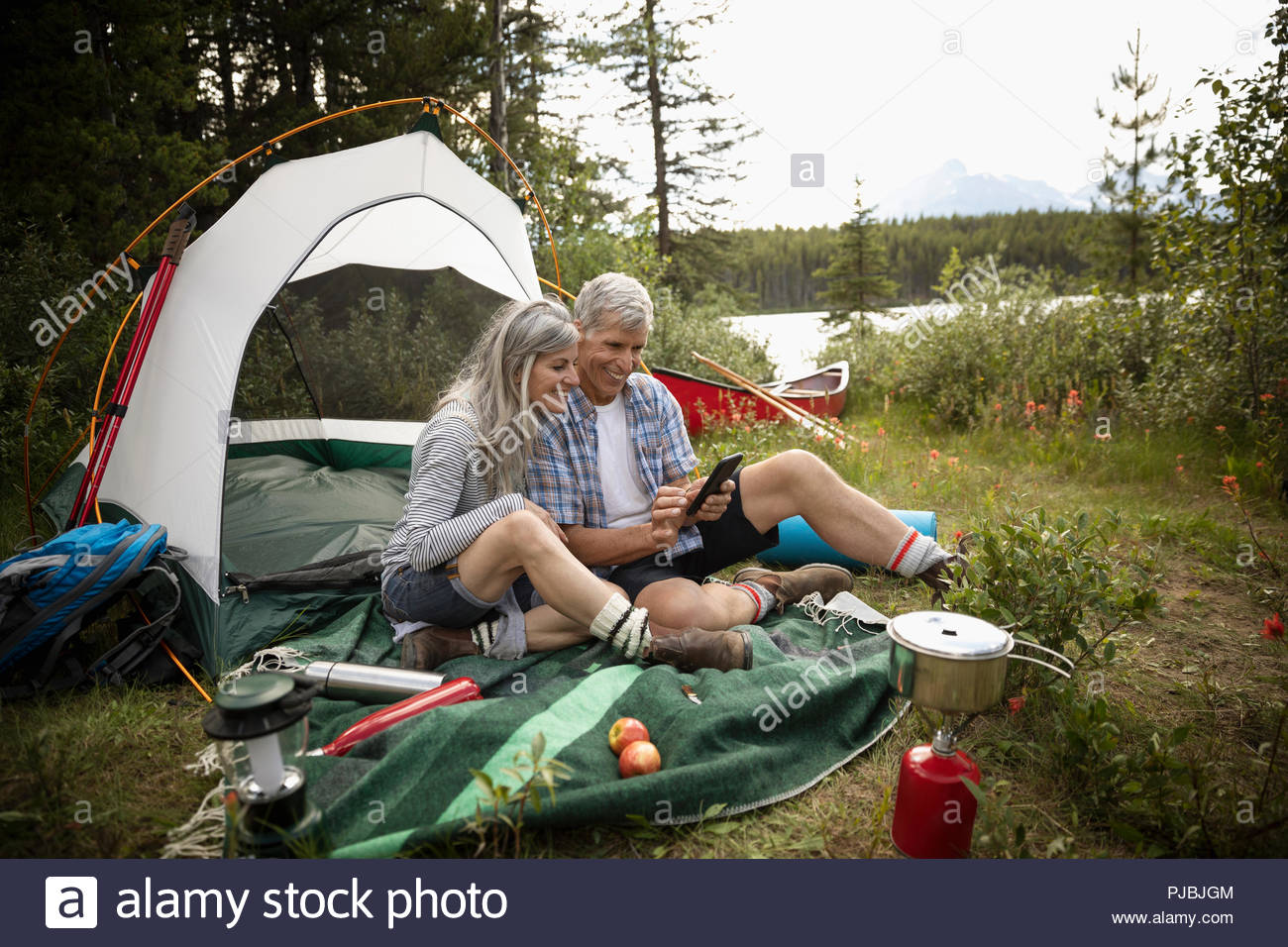 Mature couple relaxing, using digital tablet by tent at forest campsite, Alberta, Canada Stock Photo