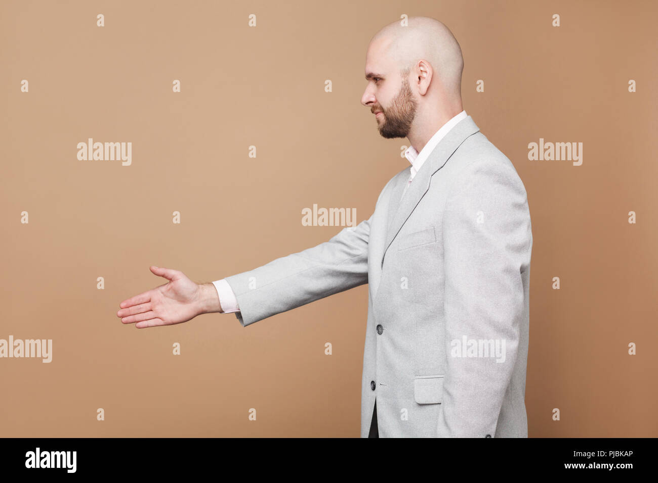 Hand shake and greeting. Profile side view portrait of middle aged bald bearded businessman in light gray suit standing and giving hand. indoor studio Stock Photo