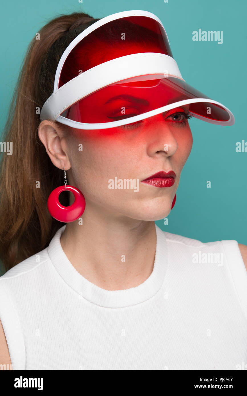 A red hair Caucasian woman in pony tail, wearing a mod 60s style earrings posing in a red visor hat, a close-up vintage style portrait. Stock Photo