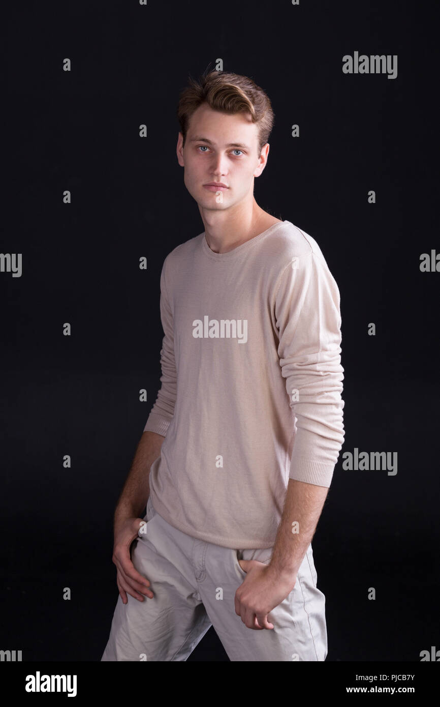 Handsome male model with blue eyes and light curlie hair in beige sweater  and white pants on a dark background Stock Photo - Alamy