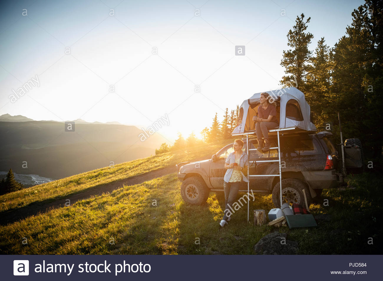 Couple camping, relaxing at SUV rooftop tent in sunny, idyllic field