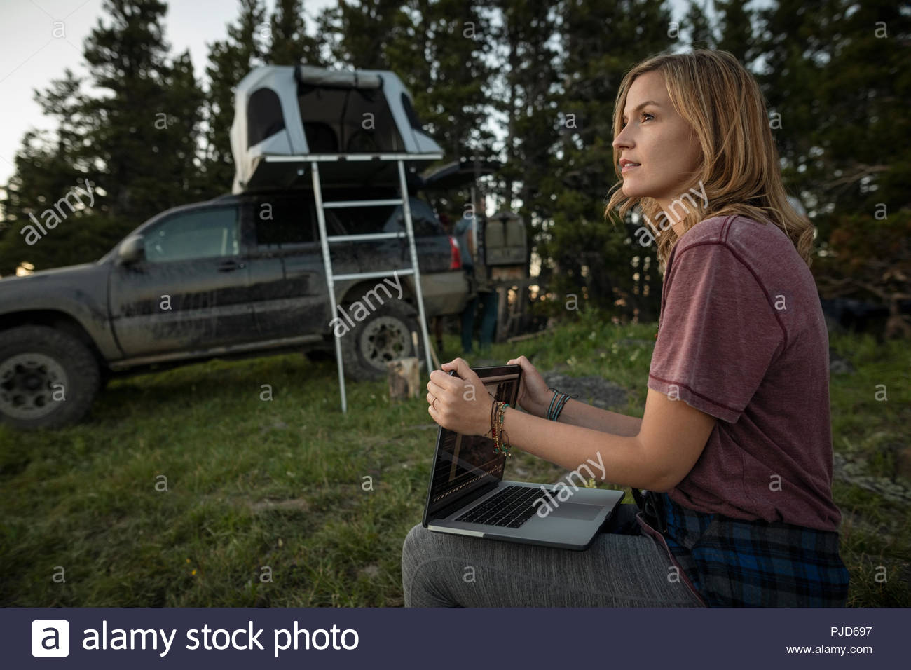 Thoughtful woman camping, using laptop near SUV with rooftop tent Stock Photo