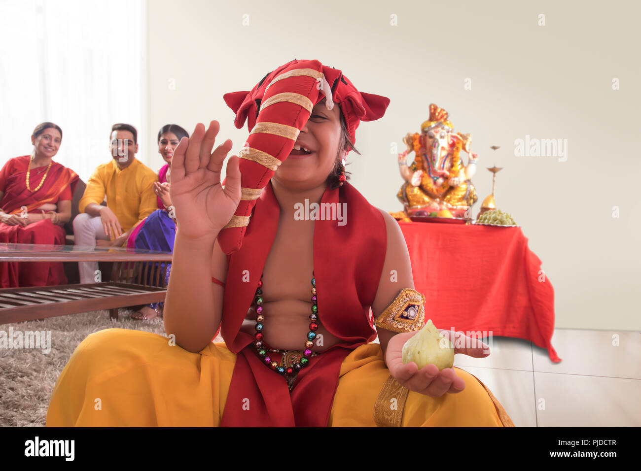 Child sitting dressed up like Ganpati with modak in one hand and Ganpati Idol in background Stock Photo