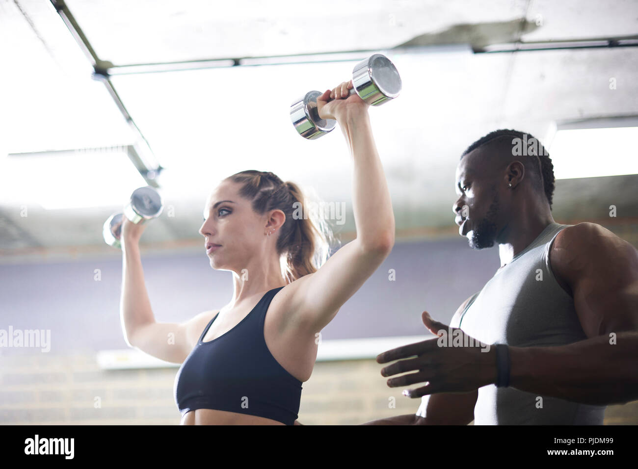 Trainer watching female client lifting dumbbells in gym Stock Photo