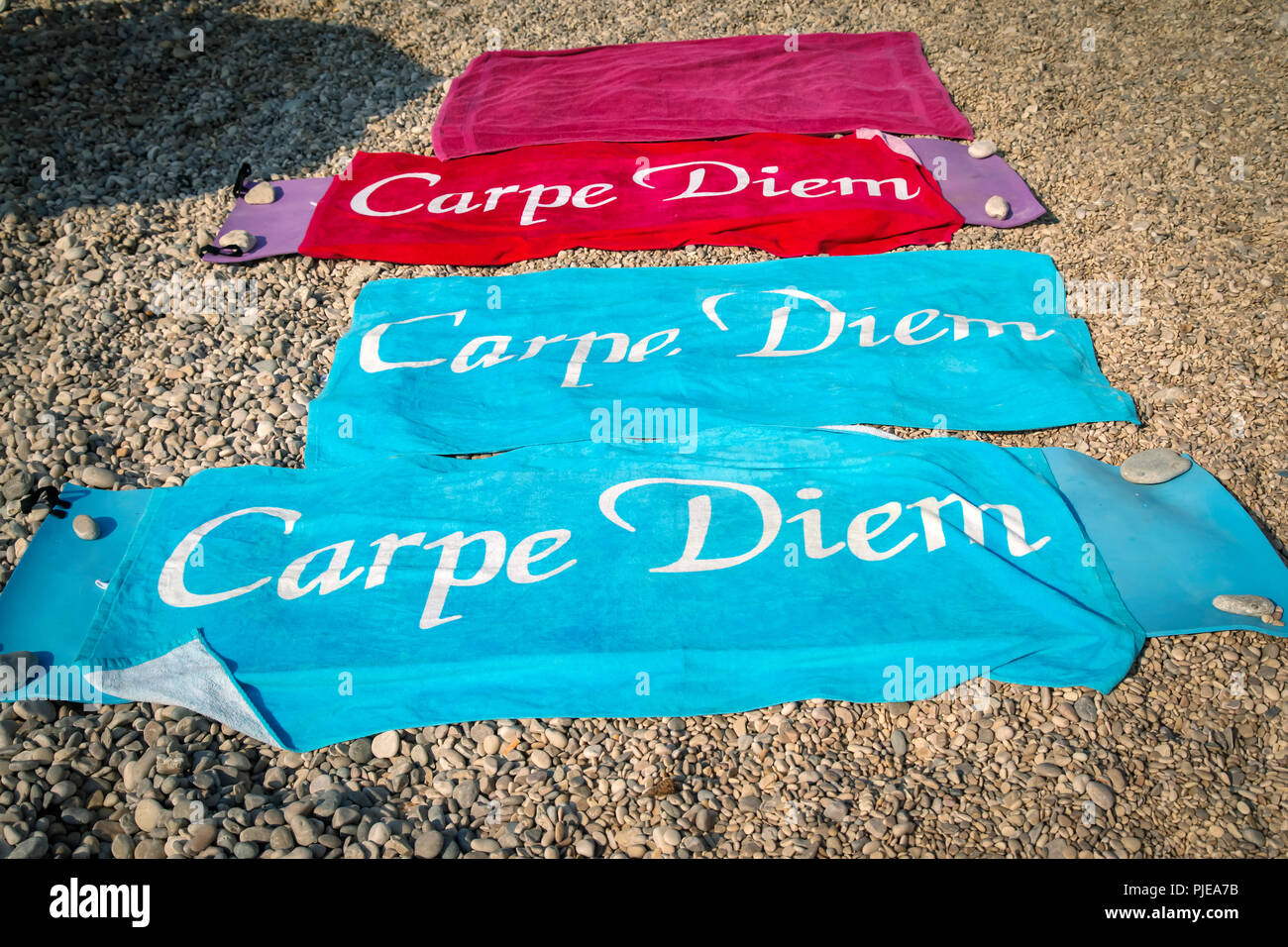 Three colorful beach towels with the inscription 'Carpe Diem', spread out on a pebble beach with rocks placed on edges to weigh them down from wind Stock Photo