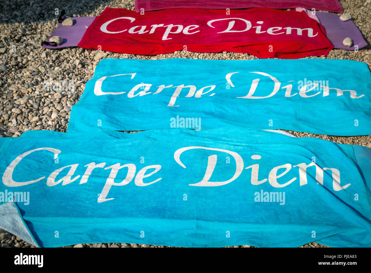 Three colorful beach towels with the inscription 'Carpe Diem', spread out on a pebble beach with rocks placed on edges to weigh them down from wind Stock Photo