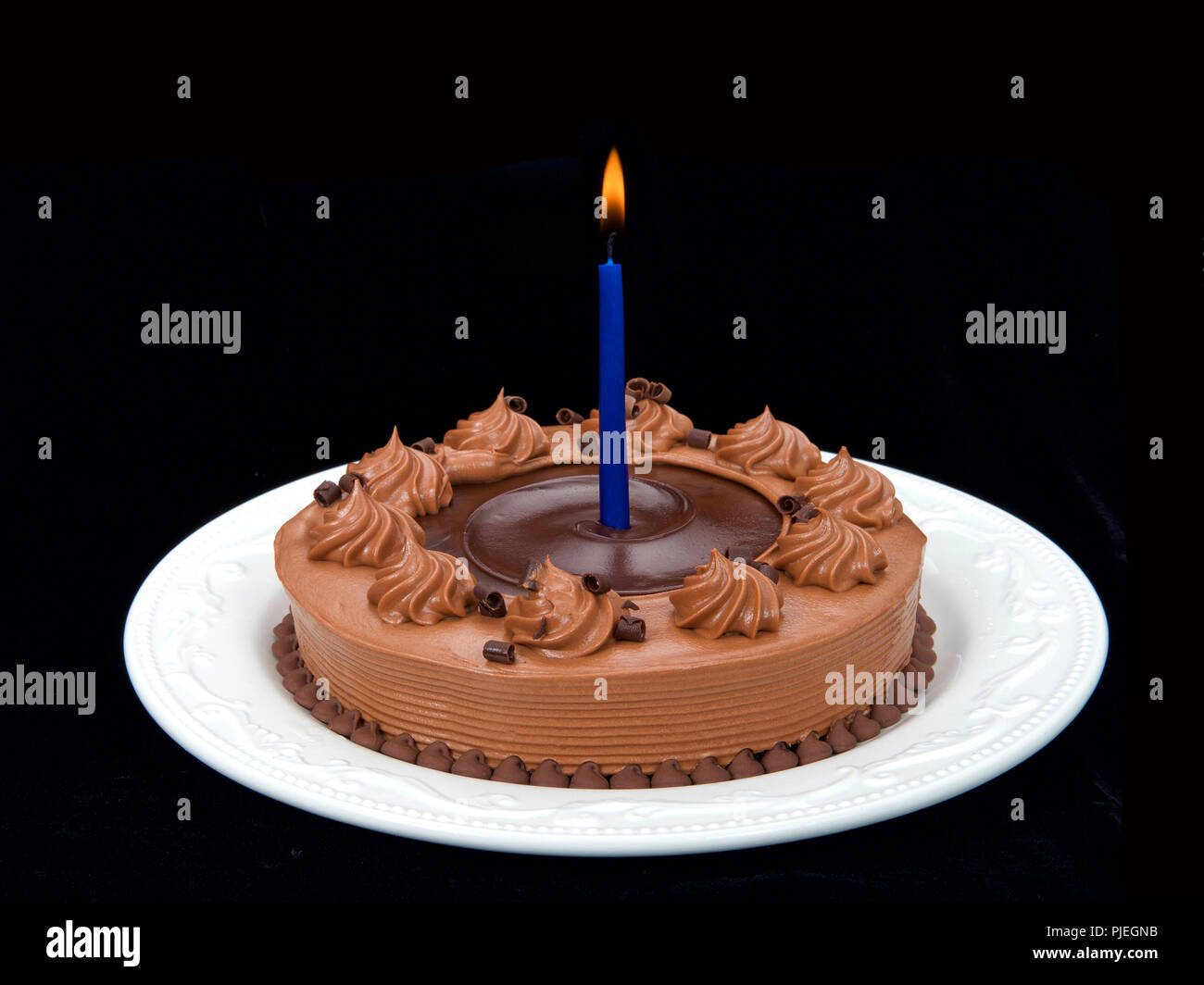 Home made chocolate cake with chocolate fudge on top on an off white porcelain plate with blue candle burning brightly on a black background Stock Photo