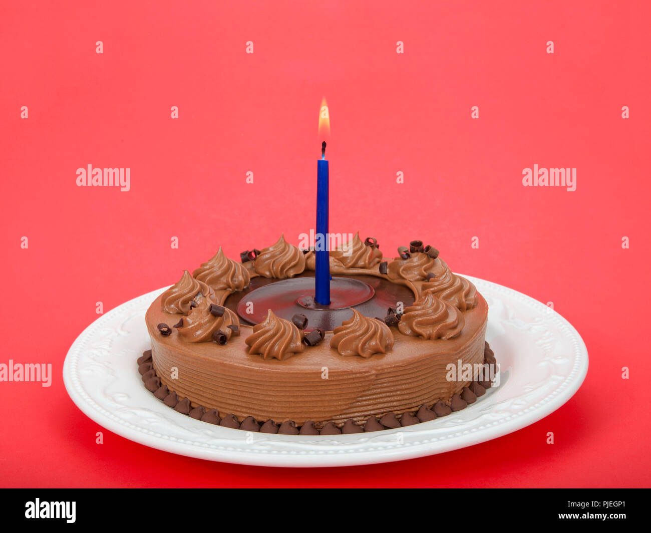 Home made chocolate cake with chocolate fudge on top on an off white porcelain plate with blue candle burning brightly on a red background Stock Photo