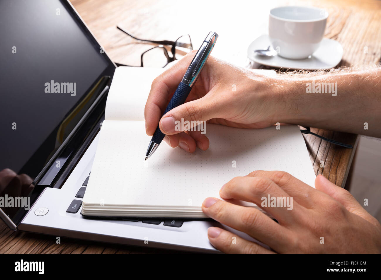 Person's Hand Writing On Black Notebook Using Pen Over Laptop Stock Photo