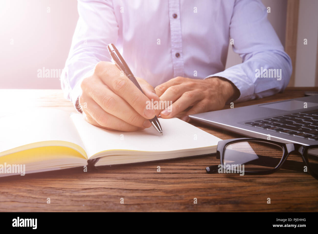 Person's Hand Writing On Black Notebook Using Pen Over Laptop Stock Photo
