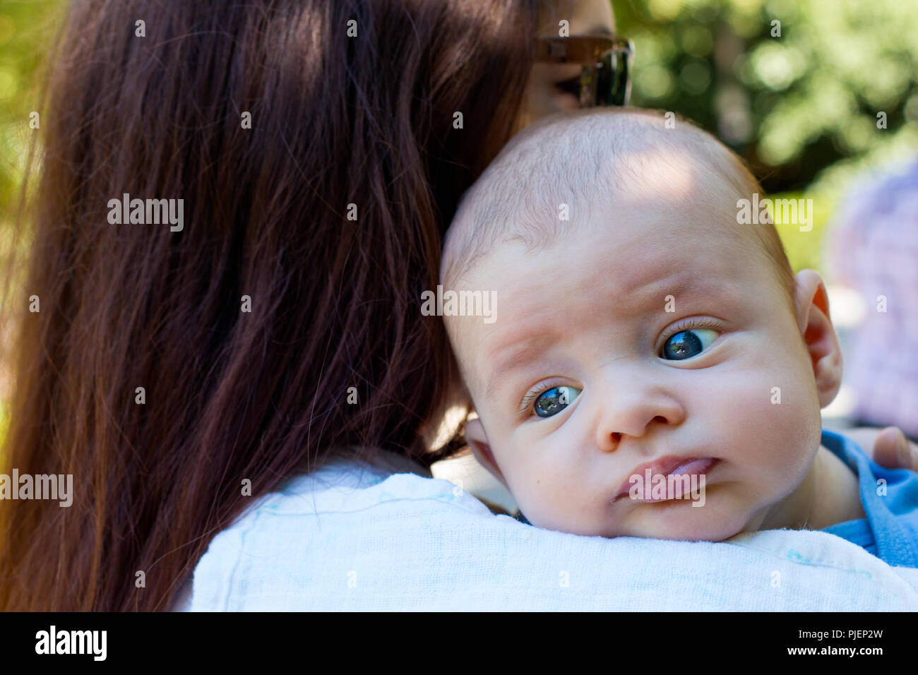 Portrait of beautiful baby with blue eyes and cute face, mother with brown hair is caring infant on her shoulder, baby burping position, day outside Stock Photo