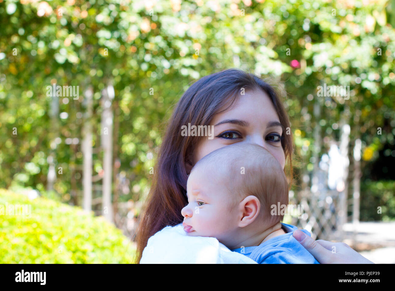 Little baby head from profile on the shoulder of young mother, pretty mom with make up on the eyes is holding baby, beautiful weather in park Stock Photo