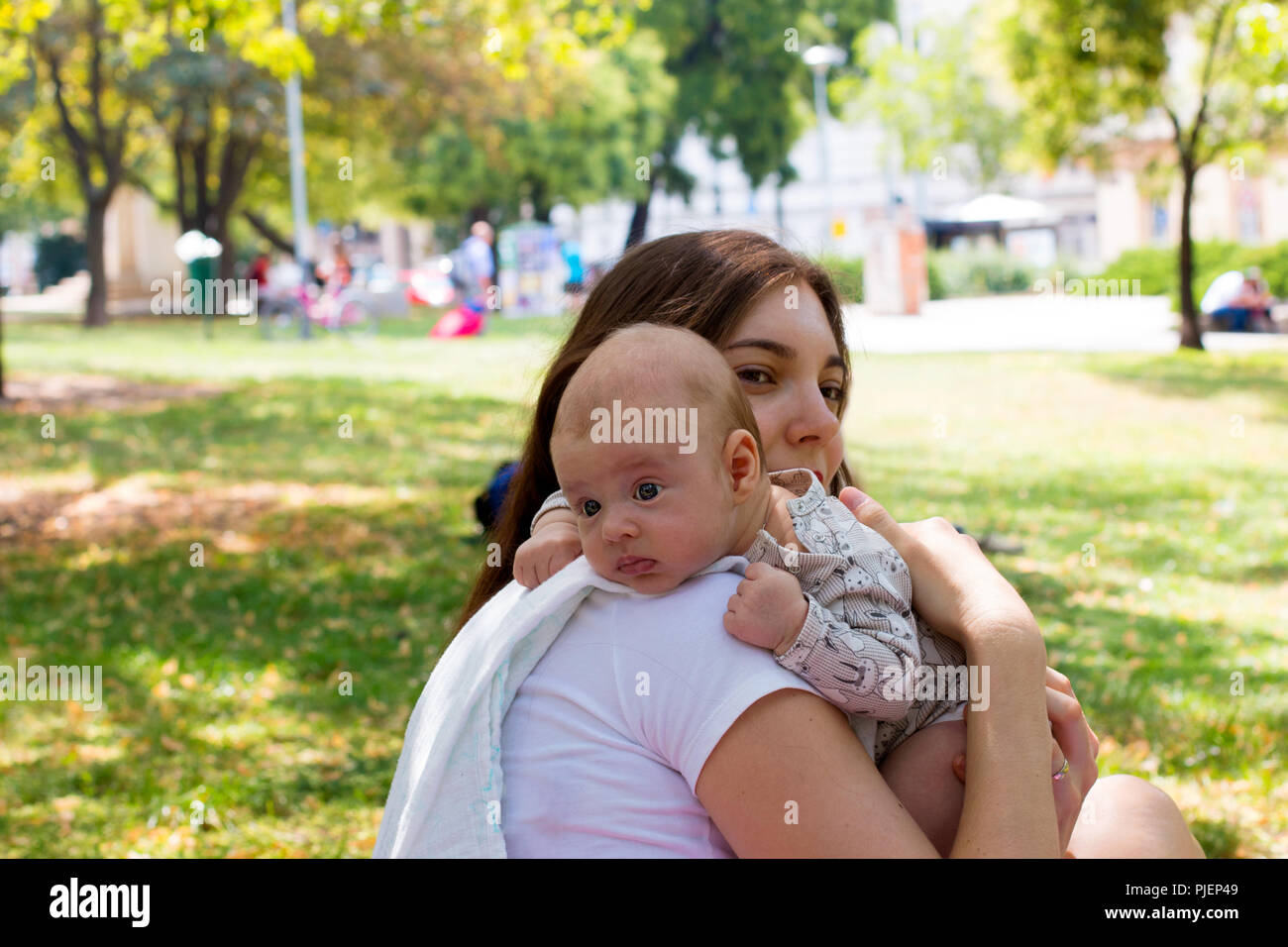 Portrait of beautiful baby resting head on mother arm, young mom is caring her infant in burping position after breastfeeding, outside in city park Stock Photo