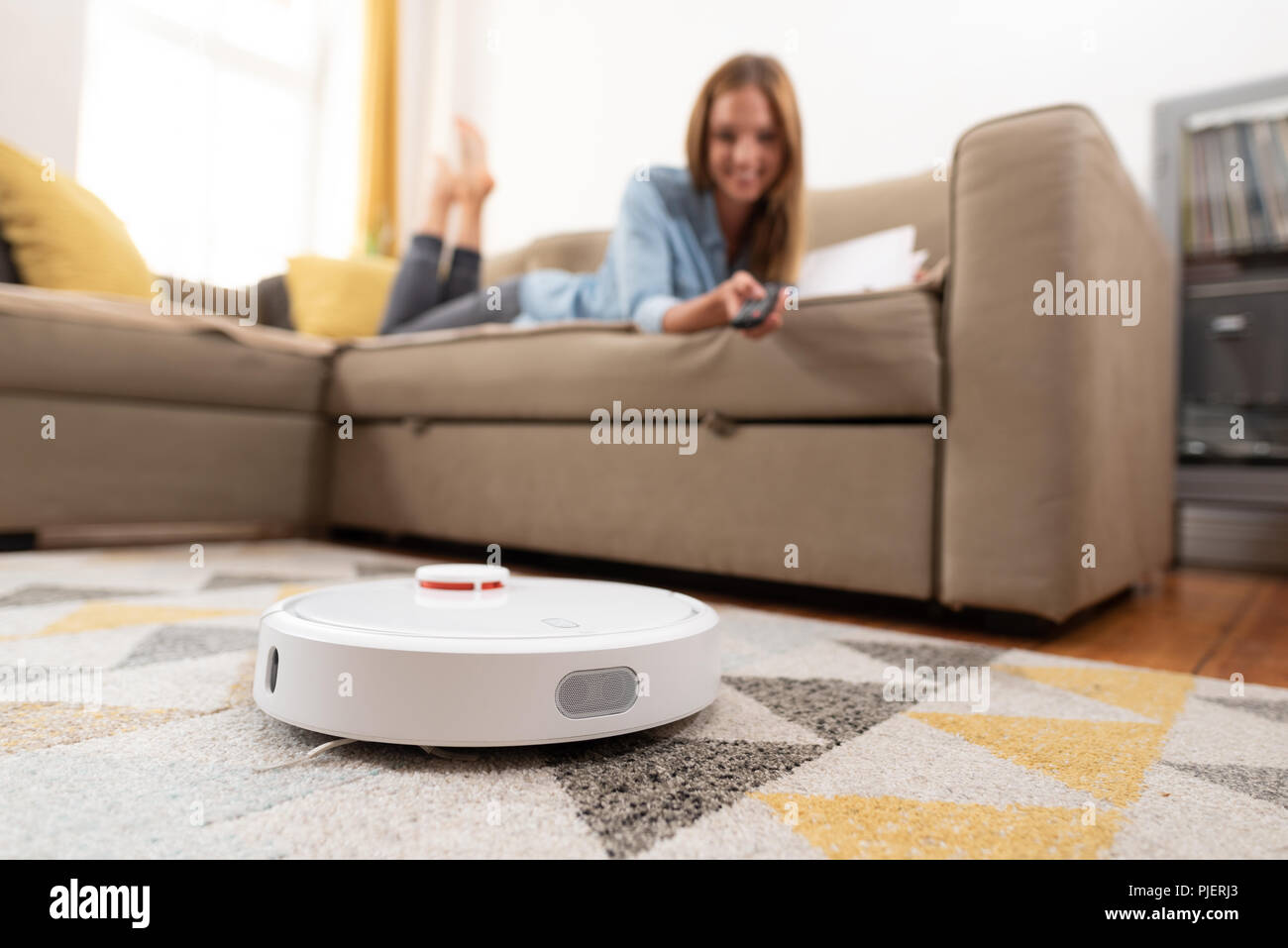 Robotic vacuum cleaner cleaning the room while woman relaxing on sofa. Woman controlling vacuum with remote control. Stock Photo