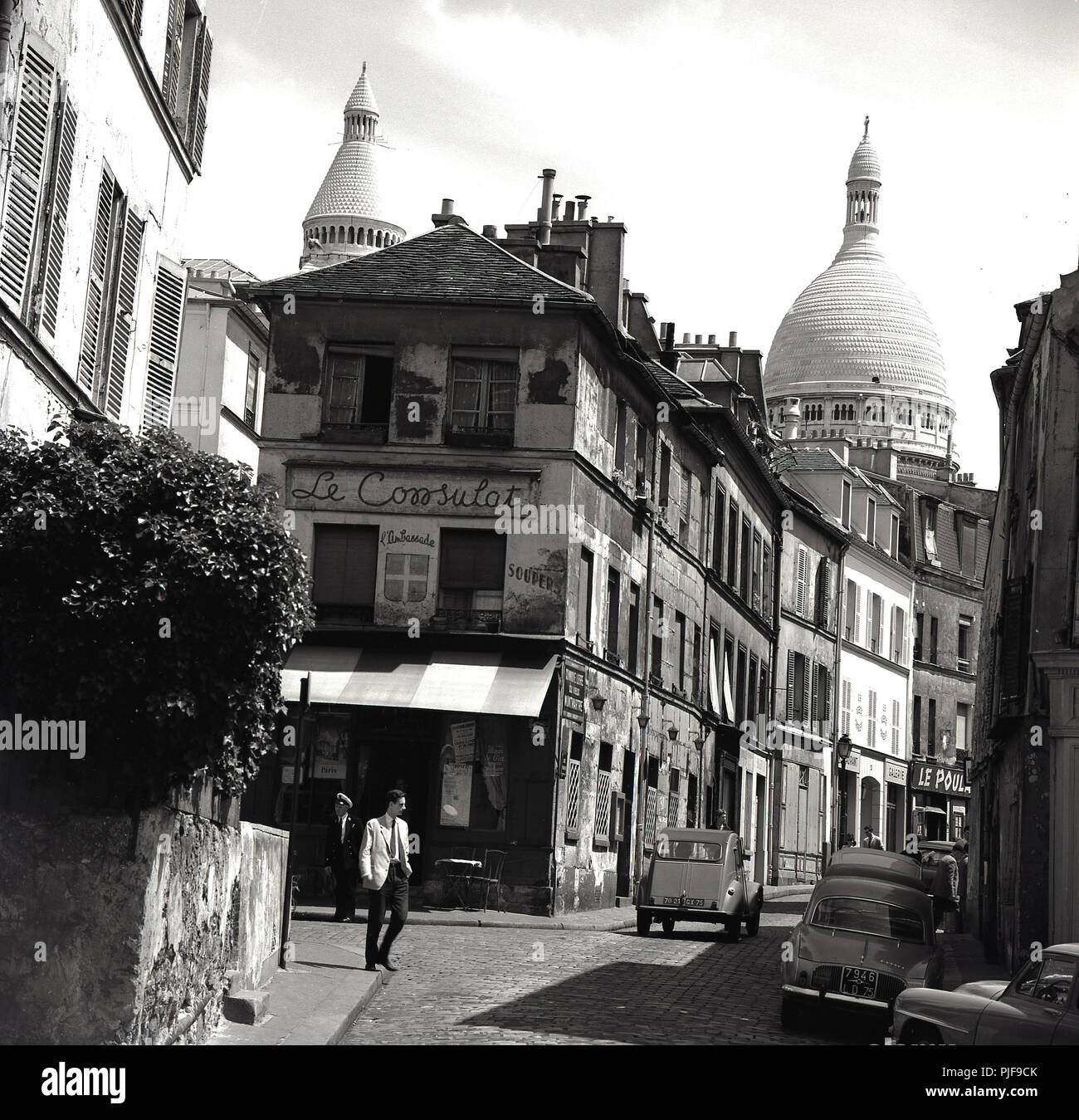 1950s, historical, a view of rue Norvins,a cobblestone street in the Montmartre district of Paris, famous for its artists, showing the decaying building housing the old Parisan cafe, Le Consulat and in the background, the domes of of the Roman Catholic church, Sacre-Coeur. The classic french car, the Citroen 2CV can be seen in the pciture. Stock Photo