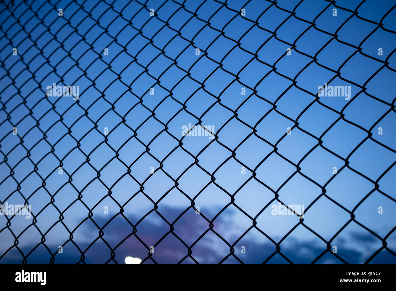Dark blue sky through wire mesh fence. Blur background, close up view of link cage, wallpaper. Stock Photo