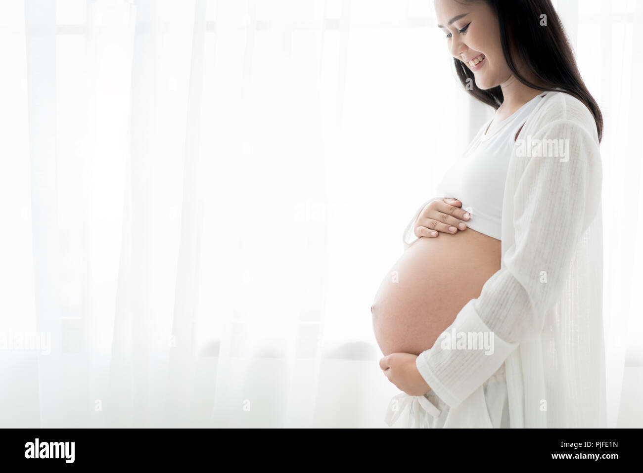 Close up of happy smiling pregnant ASian woman standingand touching her belly at home. Pregnancy, rest, people and expectation concept Stock Photo