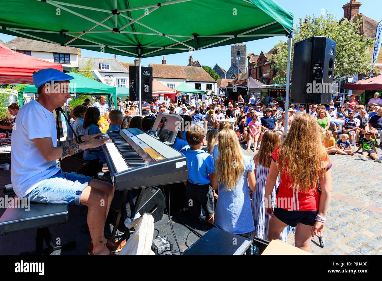 Over the shoulder view of keyboard player with school children in front of him performing to seated outdoor audience in town square in bright sunshine Stock Photo