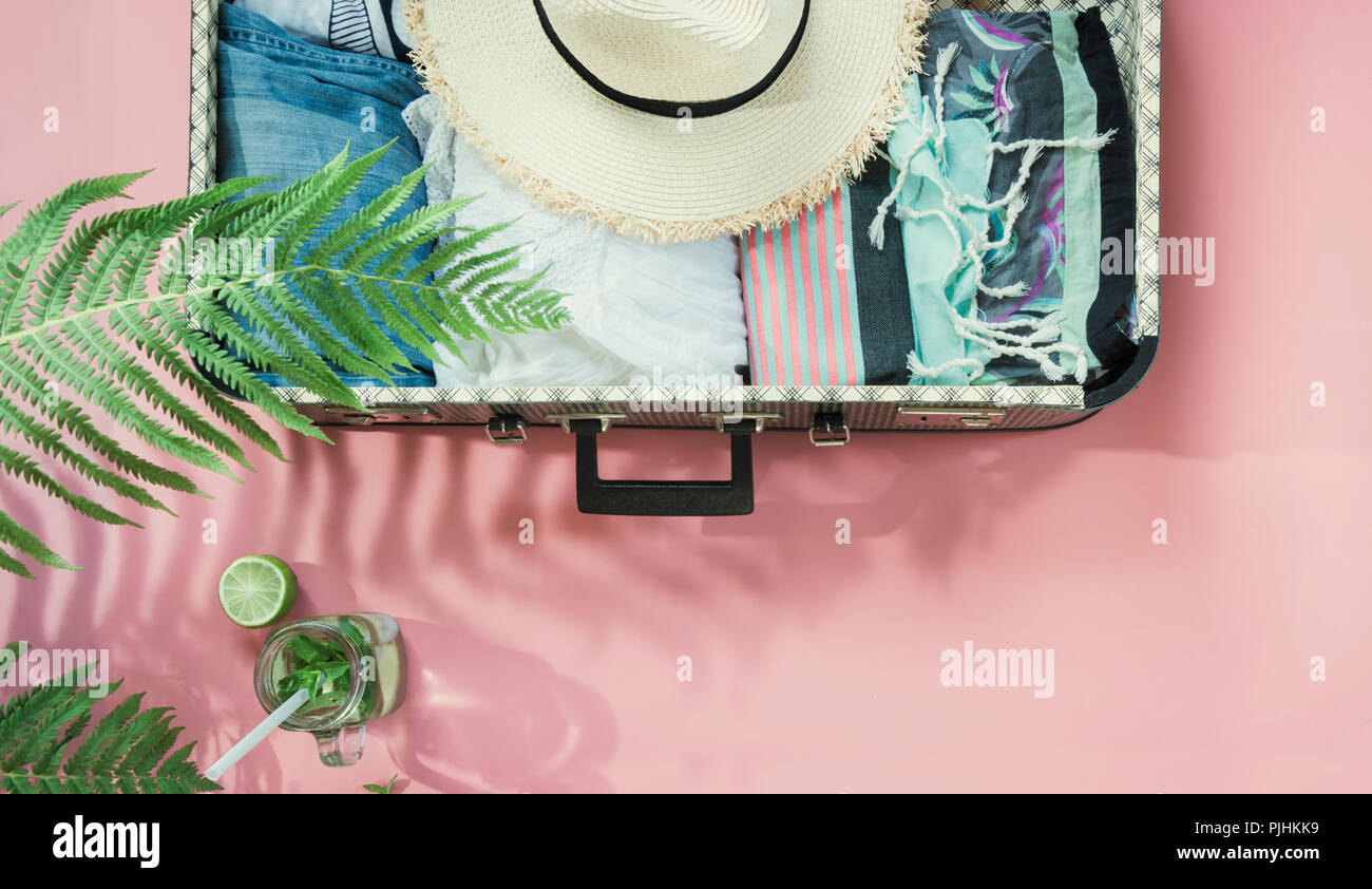 Leaf of fern, tropical detox water and open suitcase with clothes on pastel pink. Top view with copy space. Summer concept travel. Stock Photo