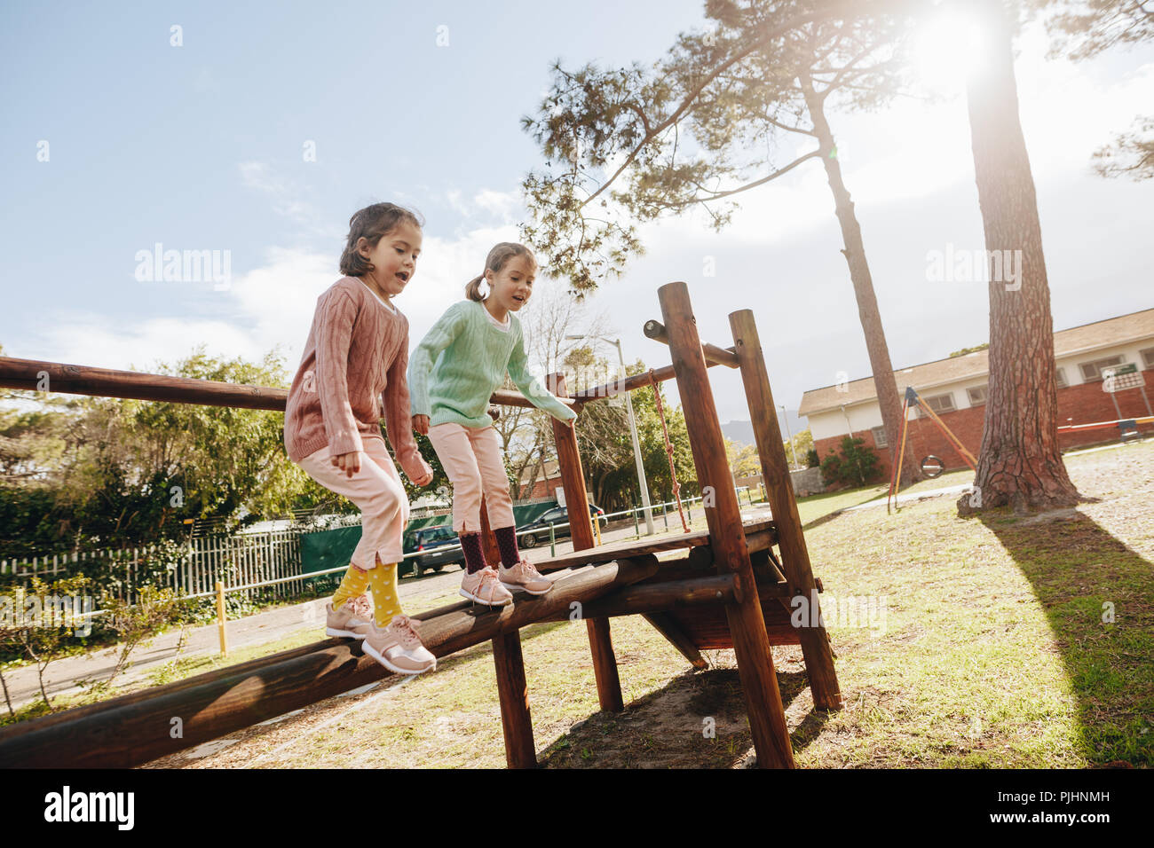 Cute little girls having fun on outdoor playground. Twin sisters jumping from wooden log at the park on sunny day. Stock Photo