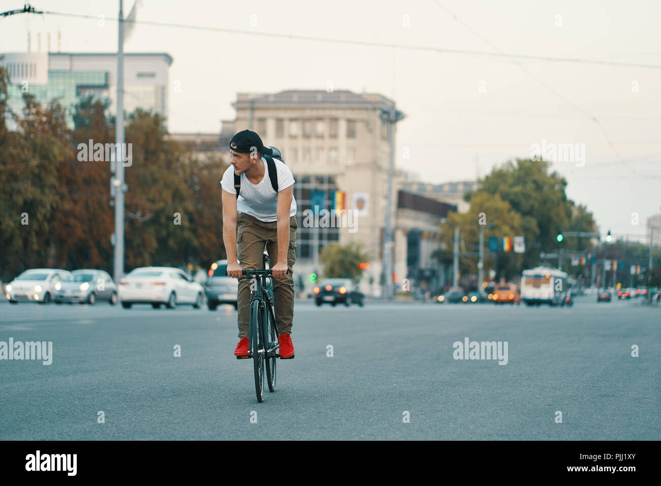 Portrait of a young man riding on bicycle in the city road, street with city far in the background. Male on black bicycle with white shirt, cap, backp Stock Photo