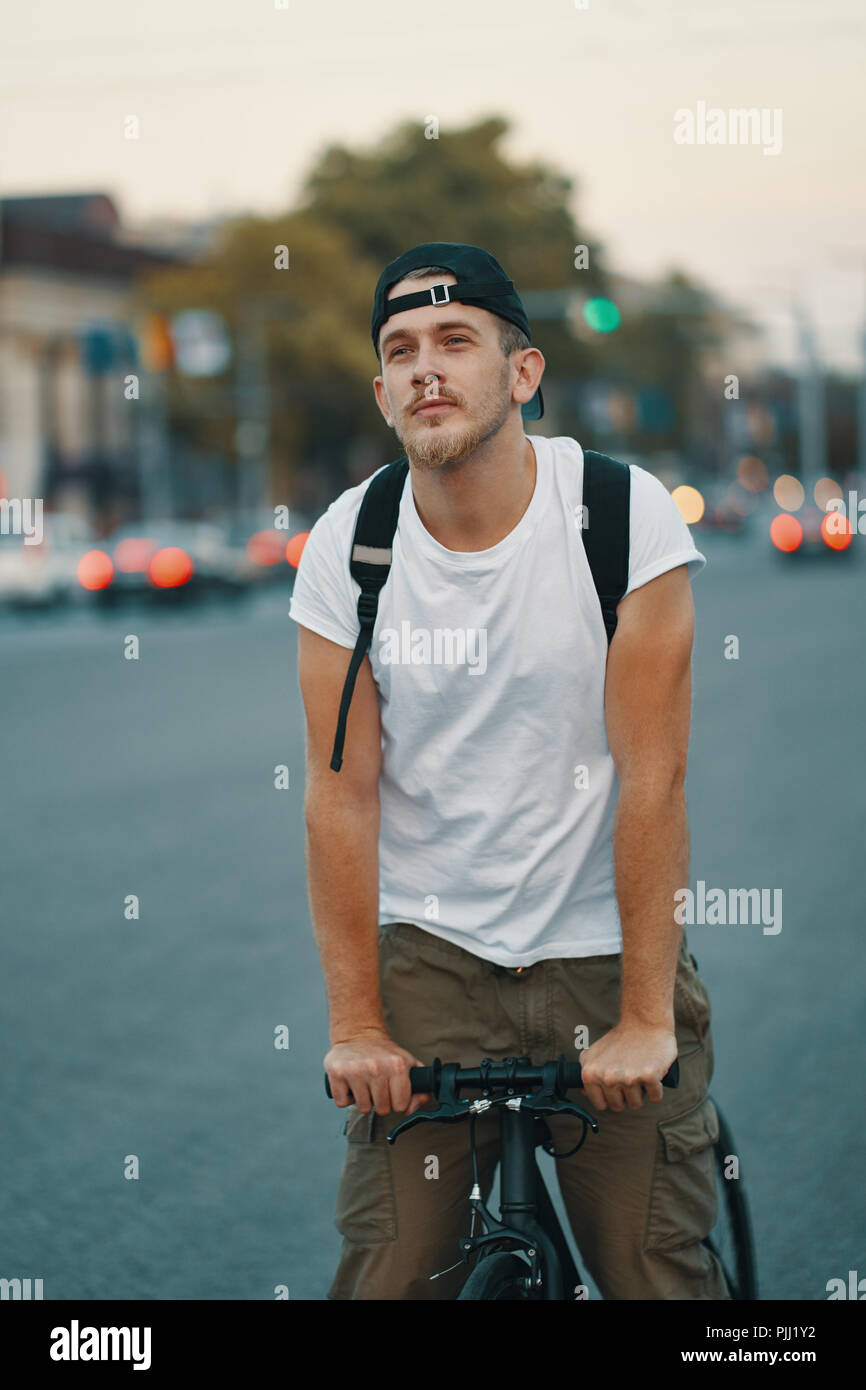 Portrait of a young man riding on bicycle in the city road, street with city far in the background. Male on black bicycle with white shirt, cap, backp Stock Photo