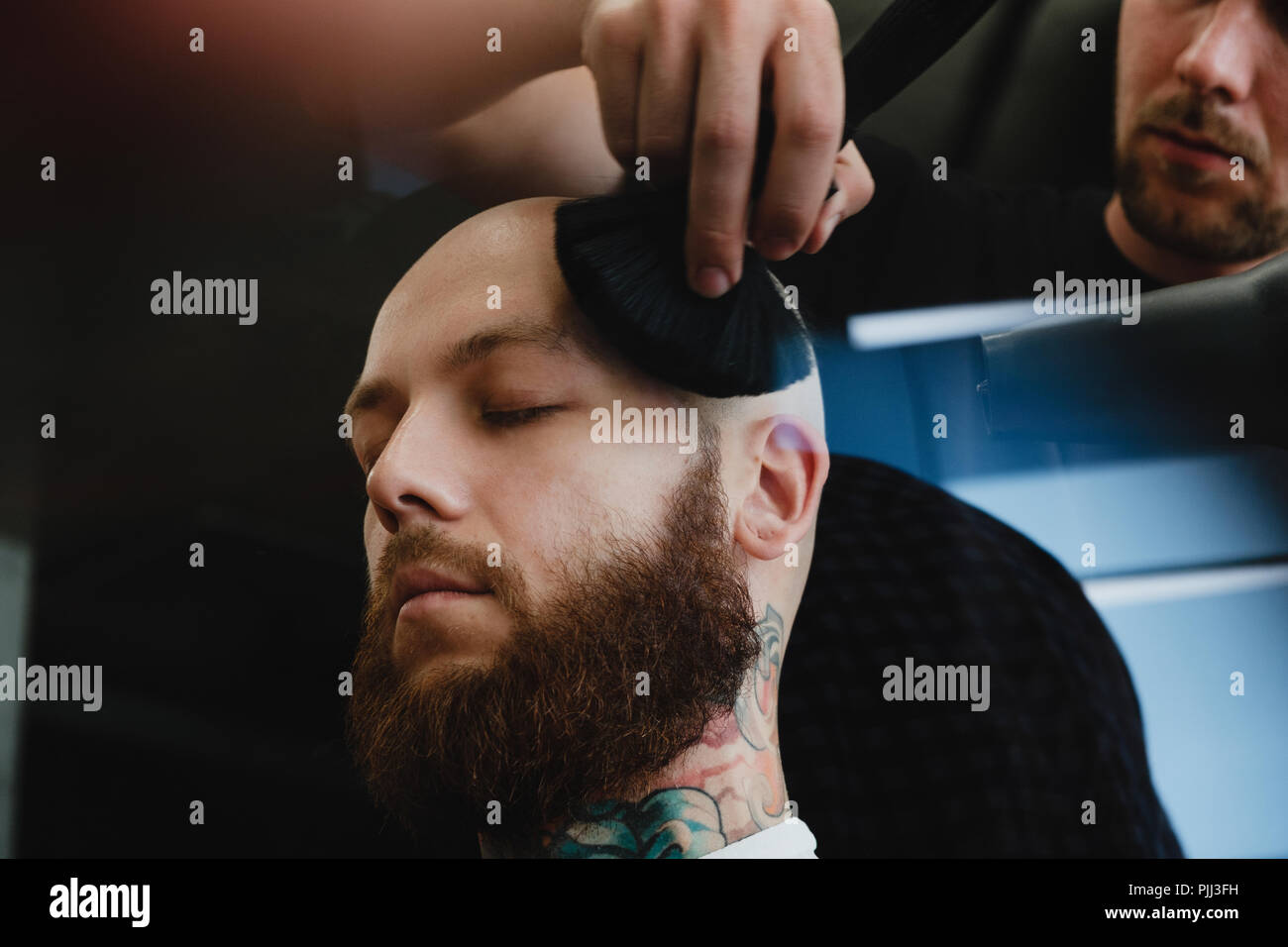 A handsome bearded skinhead man in a barbershop. The barber swipes off the cut hair with a brush. Stock Photo