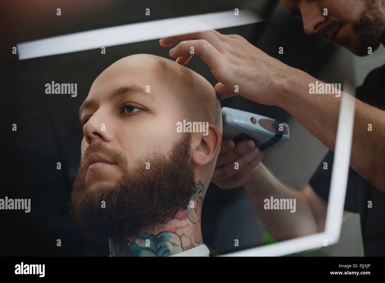 A handsome bearded skinhead man in a  barbershop. The barber shaves his head with an electric trimmer. Stock Photo