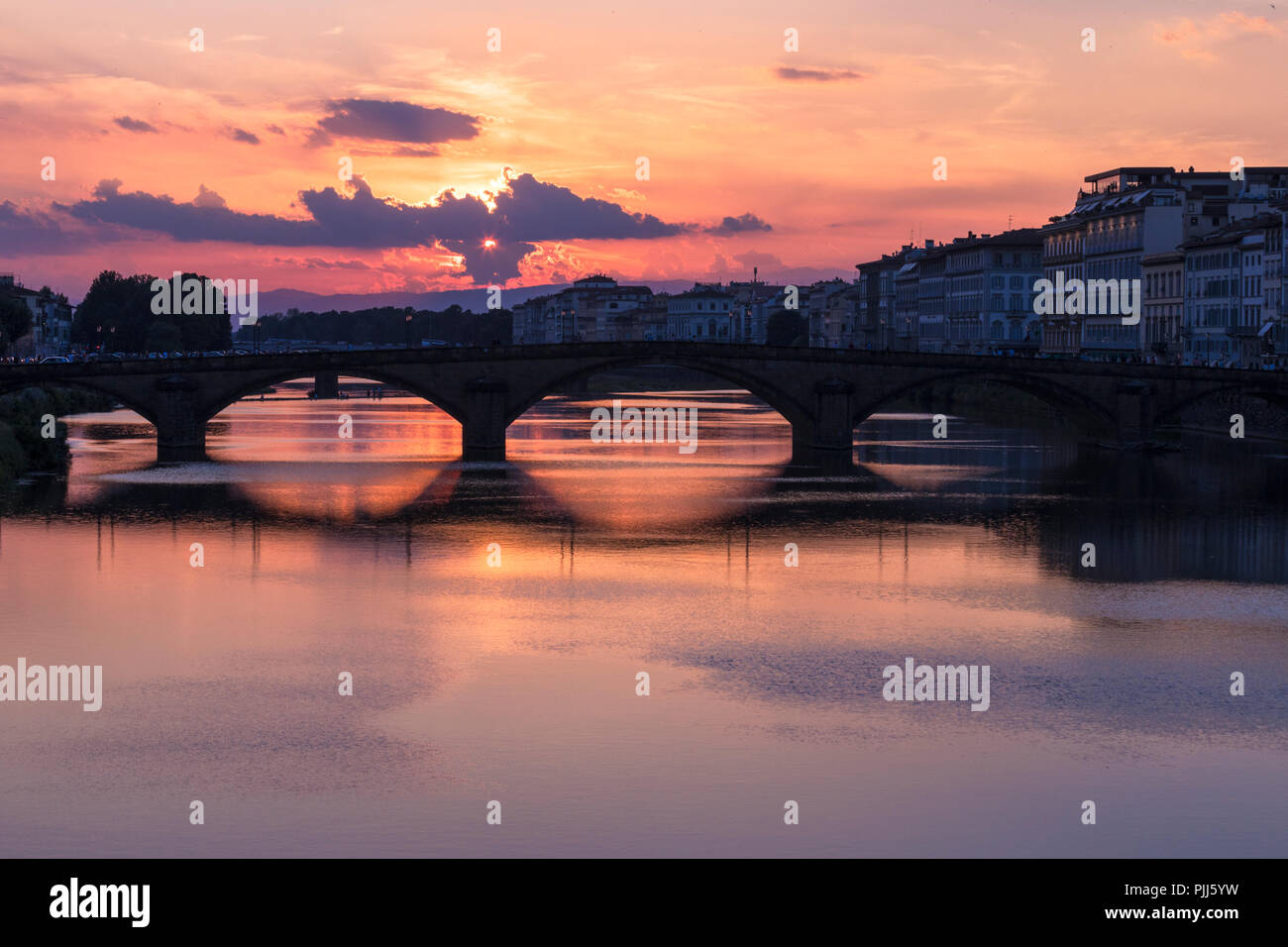 Ponte alla Carraia spans the River Arno in the city of Florence. Here the dramatic sky, reflection and stillness at sunset is captured. Stock Photo