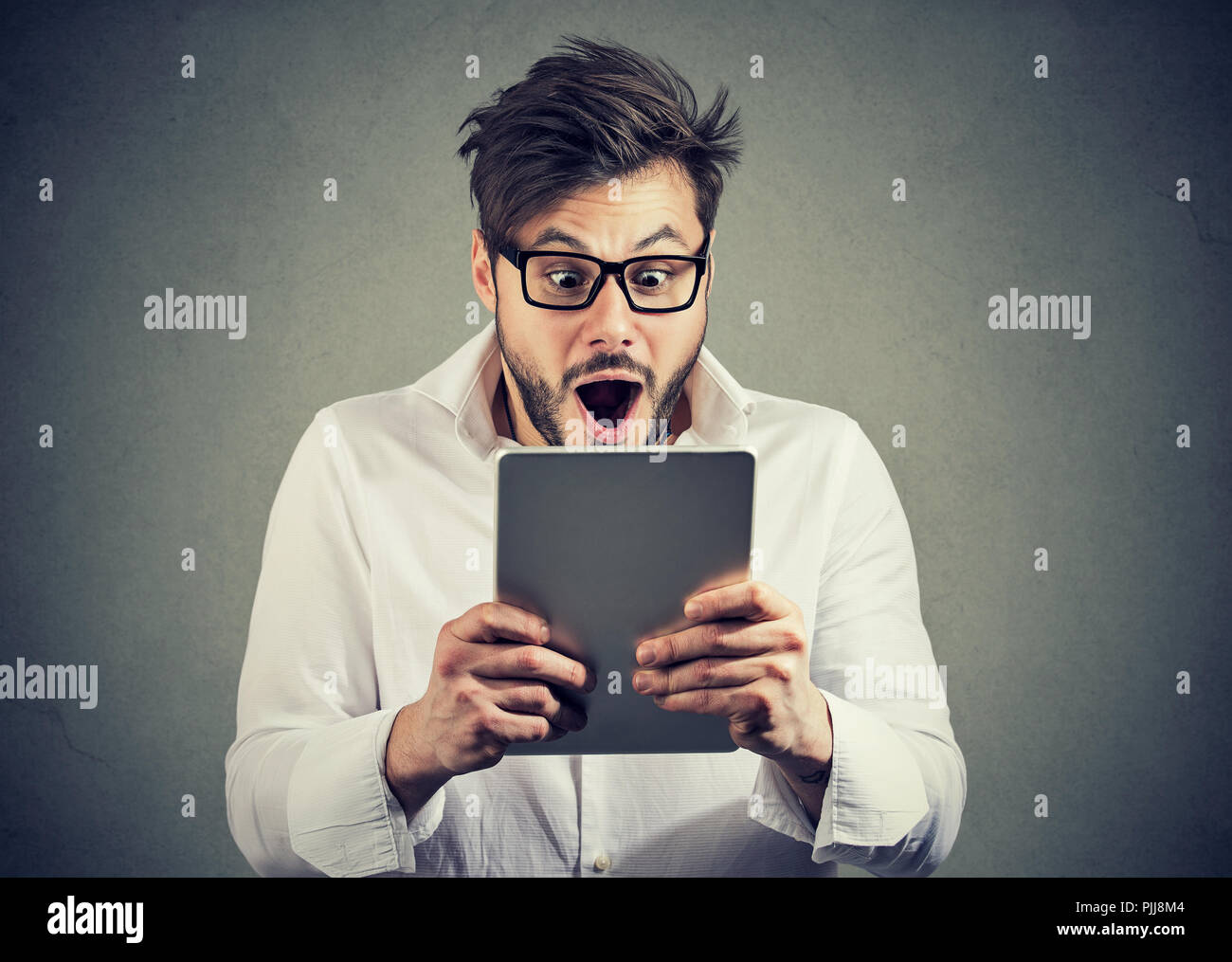 Young man in eyeglasses watching tablet with shock having bad news and posing on gray background Stock Photo