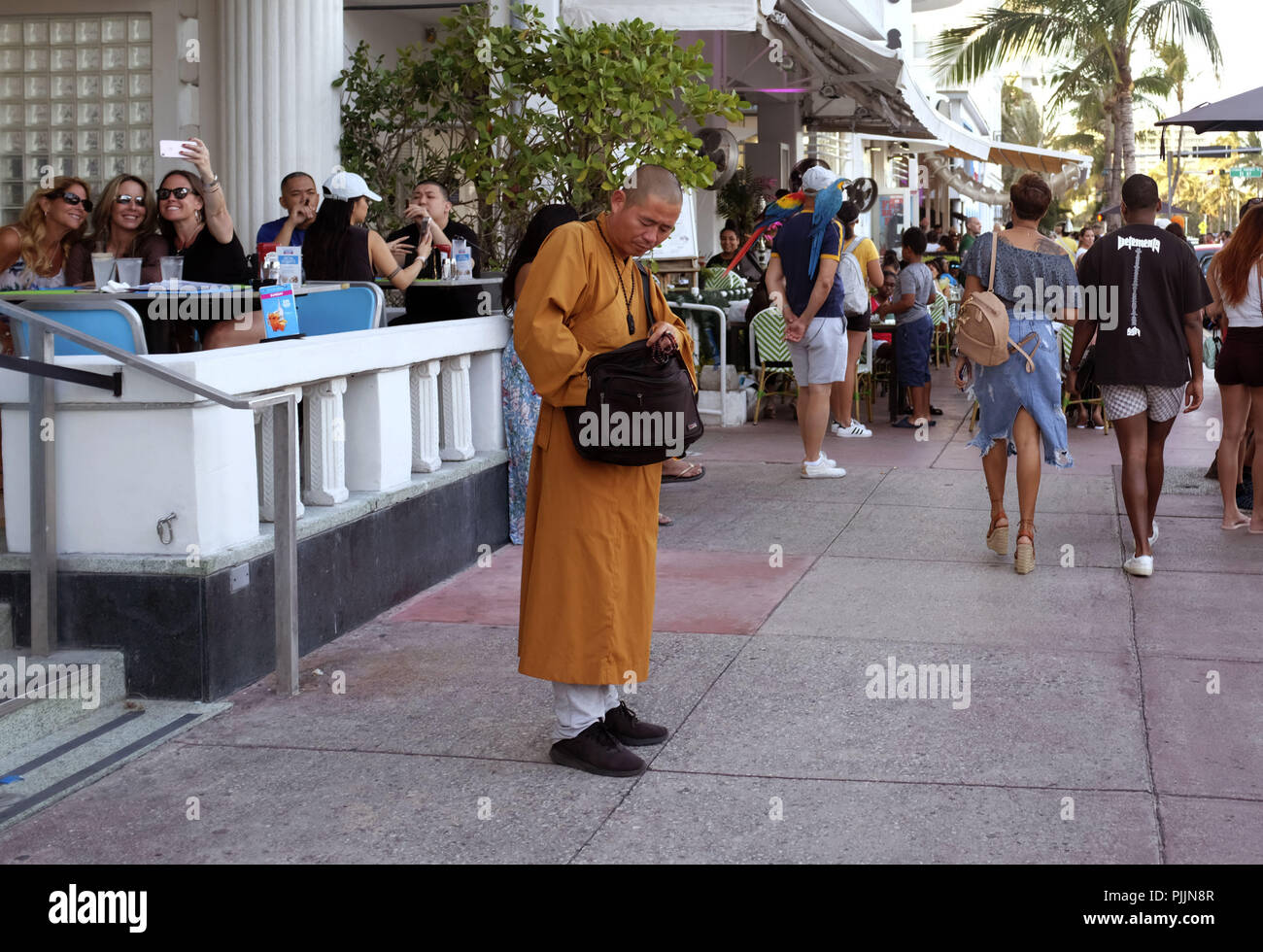 Miami, Florida, USA. 25th Feb, 2018. A monk stands alone in the middle of a South Beach, Miami sidewalk holding his prayer beads. He is lost in a sea of crazyness that surrounds South Beach. A man stands in the background with macaw birds on his shoulders, while women off to the side take selfies. Credit: Allison Dinner/ZUMA Wire/Alamy Live News Stock Photo